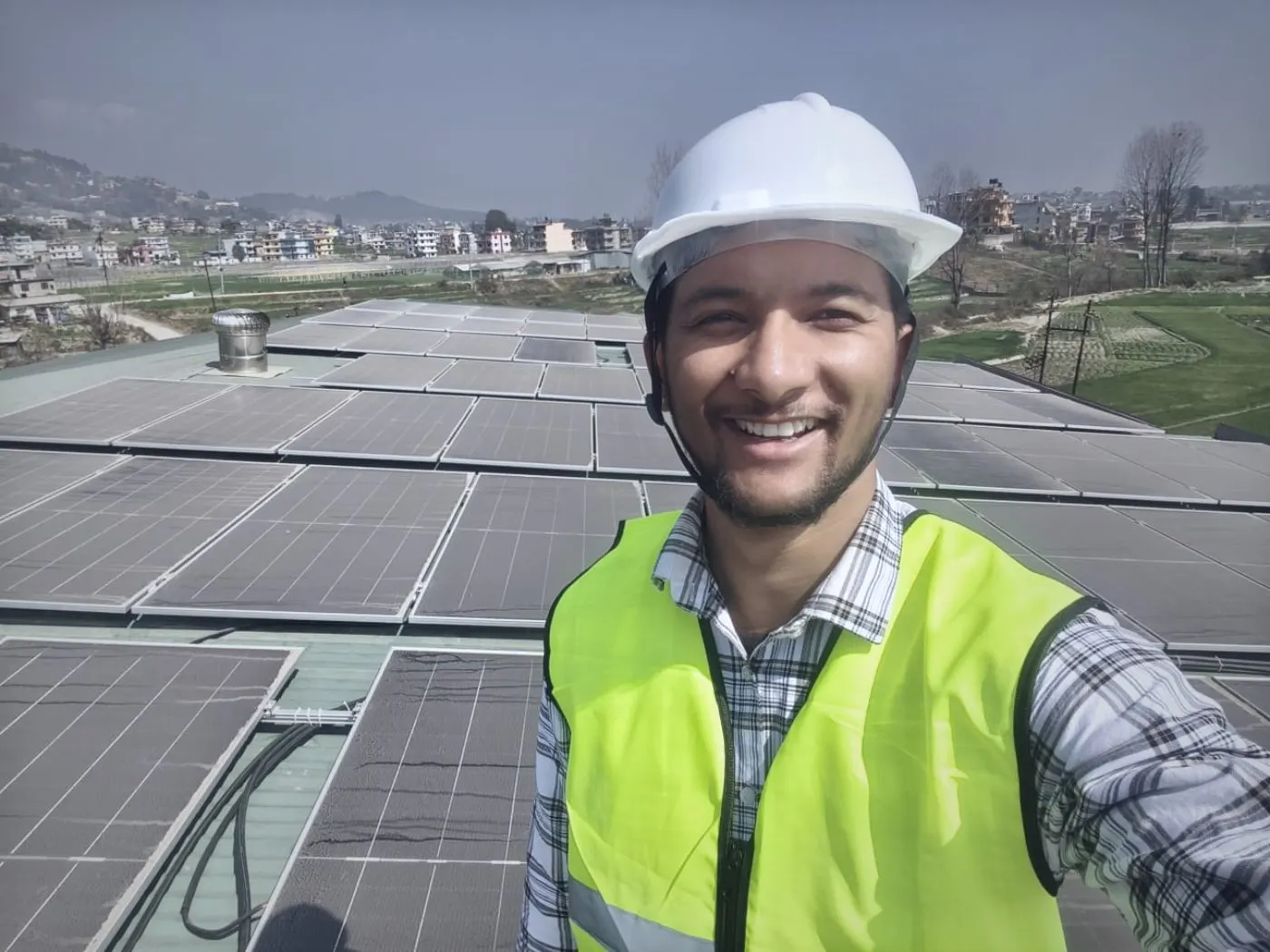 Standing on a commercial rooftop array in the Kathmandu valley, hard hat and hi-vis vest on, with rows of modules and the valley's terraced fields and settlements in the background